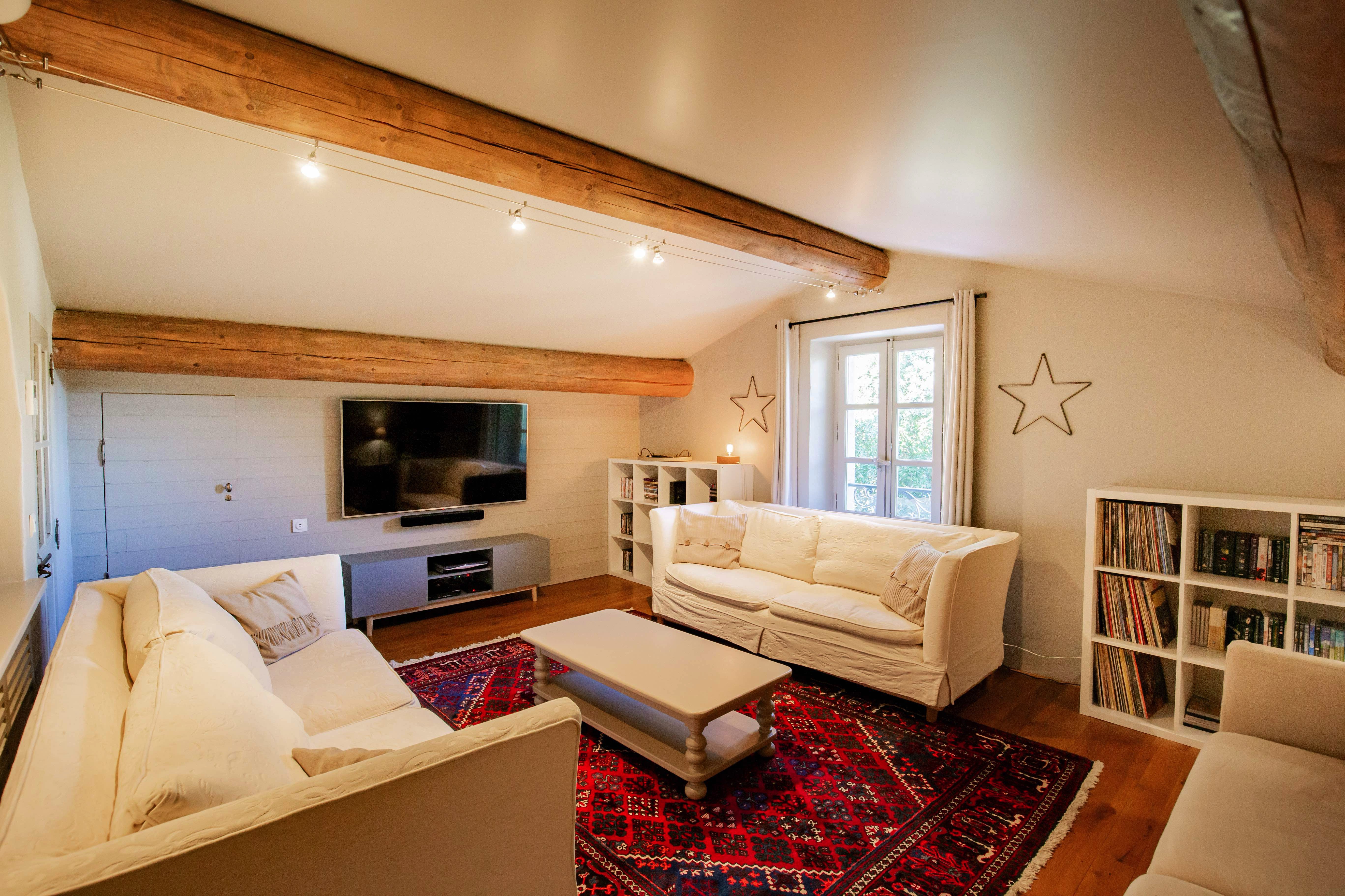 Cozy living room with white sofas, a red patterned rug, and wooden beams across the ceiling. there's a tv, bookshelves, and decorative stars on the walls, with natural light coming from a window.